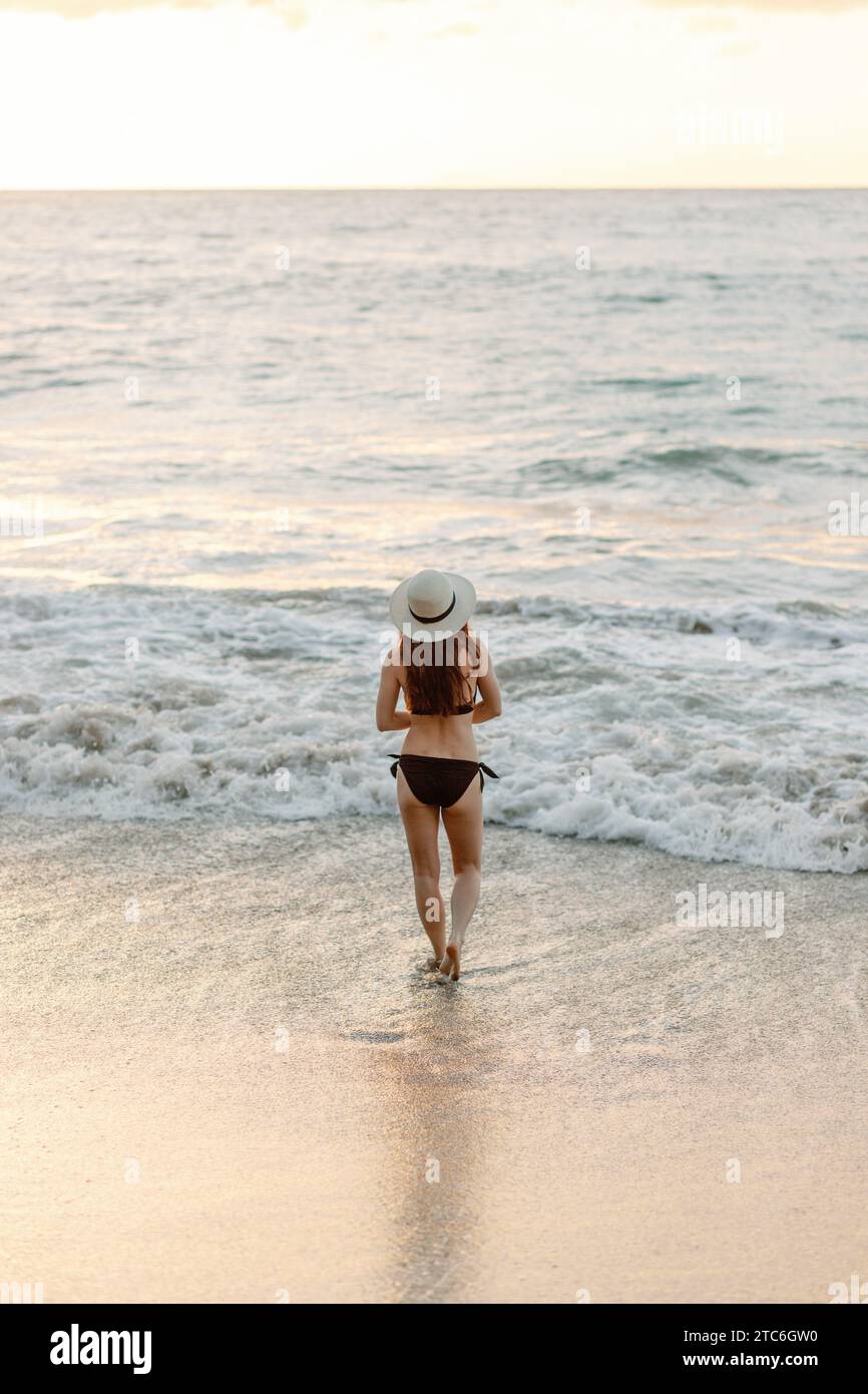 Besinnliche junge Frau in schwarzem Bikini geht bei Sonnenuntergang am Strand. Stockfoto