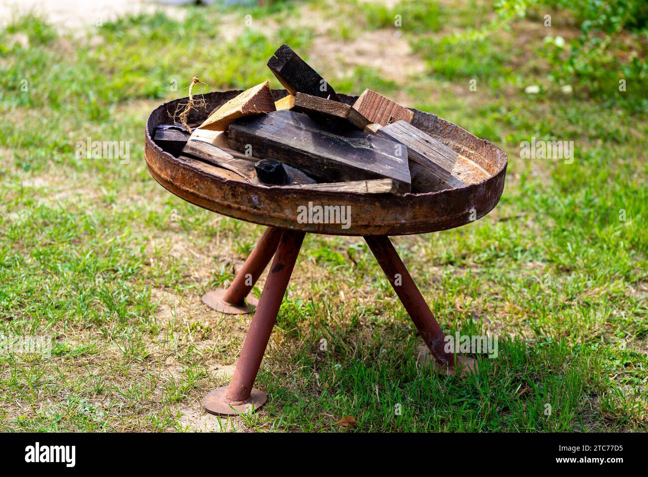 Eine rostige Feuerschüssel gefüllt mit verschiedenen Holzstücken, mit drei langen Füßen und sehr krumm auf dem Gras Stockfoto