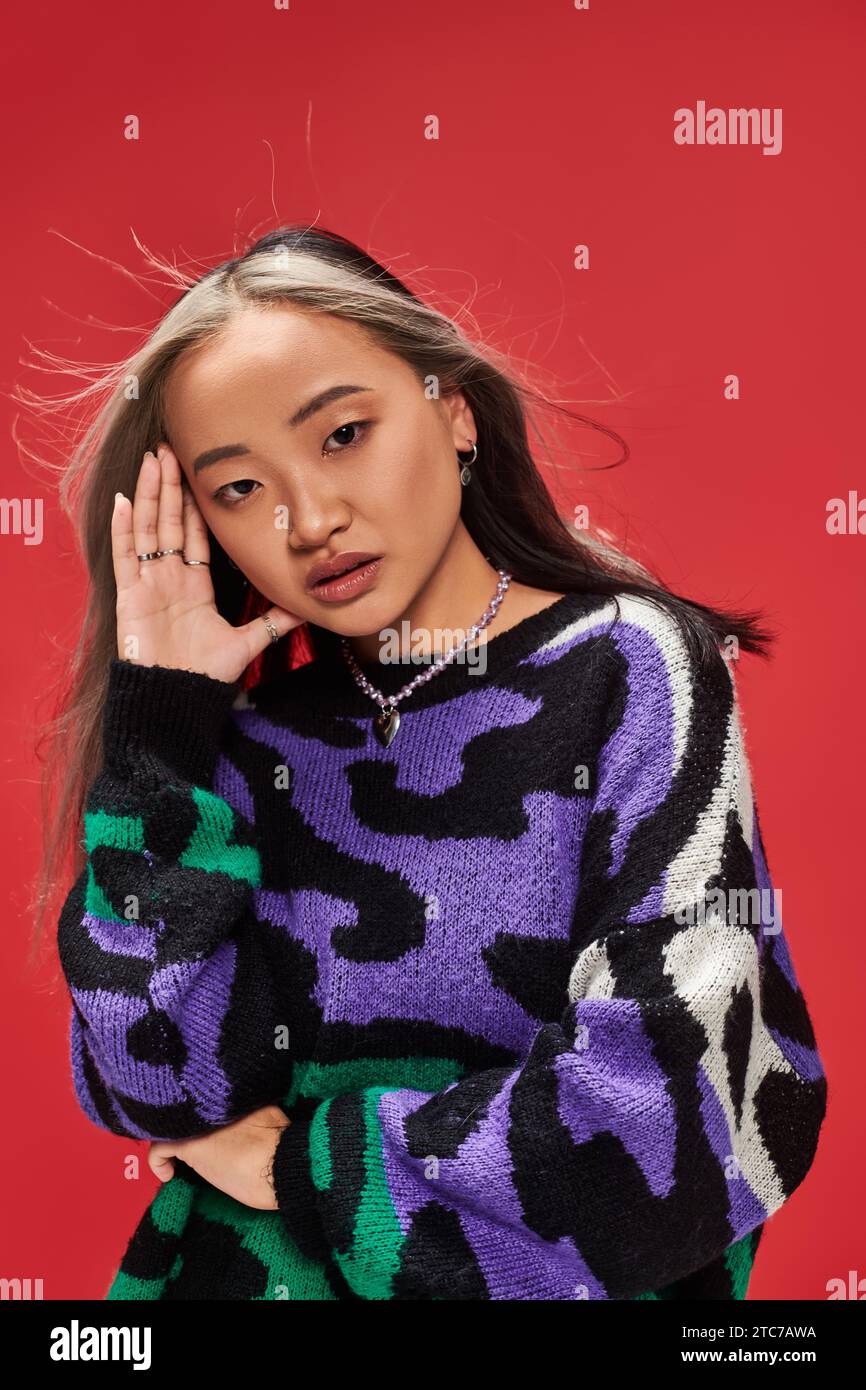 Verführerische junge asiatische Frau mit gefärbten Haaren im Pullover mit Tierdruck, die mit der Hand in der Nähe des Gesichts posiert Stockfoto