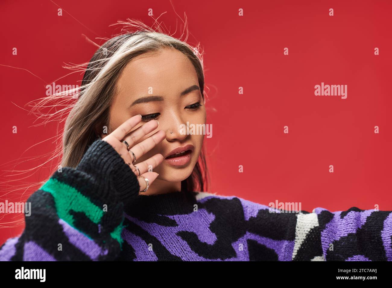 Junge asiatische Frau mit gefärbten Haaren im Pullover mit Tierdruck posiert mit Hand in Gesichtsnähe, Wind Stockfoto
