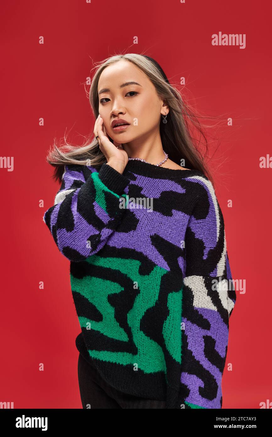 Hübsche junge asiatische Frau mit gefärbten Haaren im Pullover mit Tierdruck posiert mit Hand nahe Gesicht Stockfoto