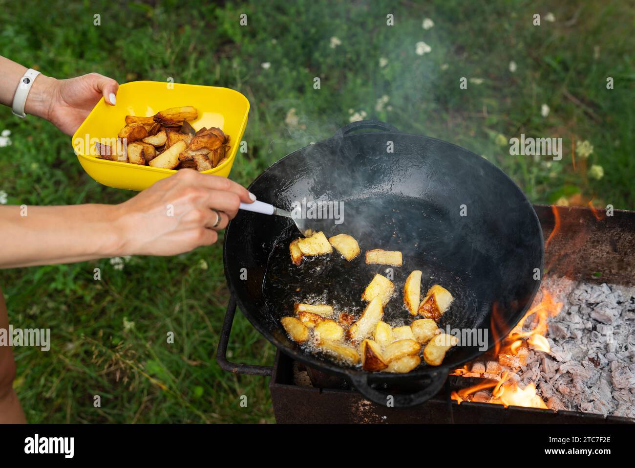 Ein Mann kocht Kartoffeln im Wok über offenem Feuer. Die fertigen Kartoffeln in eine gelbe Schüssel geben. Der Hintergrund ist ein grasbewachsenes Feld. Picknick im Freien Stockfoto