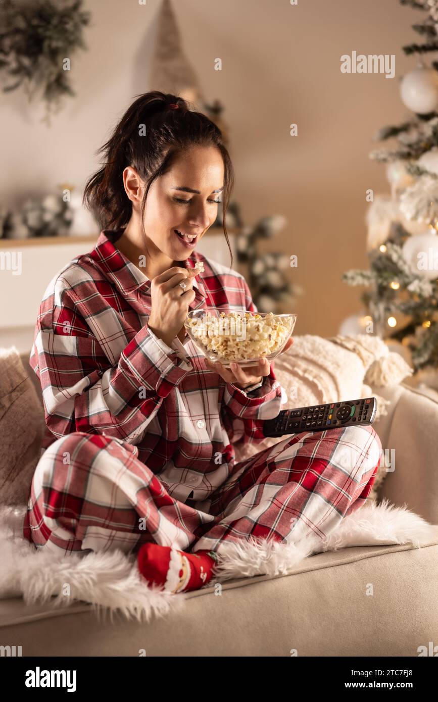 Eine junge Brünette genießt während Weihnachten Popcorn und sitzt in ihrem Pyjama auf der Couch vor dem Fernseher. Stockfoto