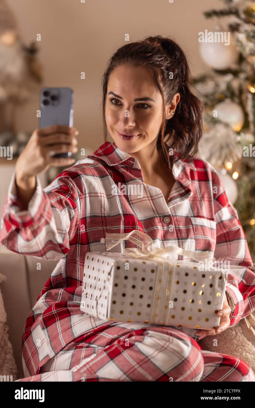 Fröhliche Frau im Pyjama sitzt auf Sofa mit Geschenk neben dem Weihnachtsbaum und nimmt Selfie. Stockfoto
