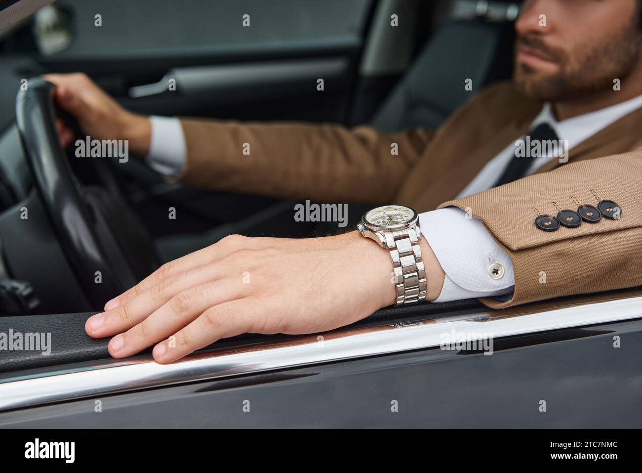 Zugeschnittene Ansicht eines selbstbewussten Geschäftsmannes in beigefarbenem Anzug und Armbanduhr, der ein Luxusauto auf der Straße fährt Stockfoto