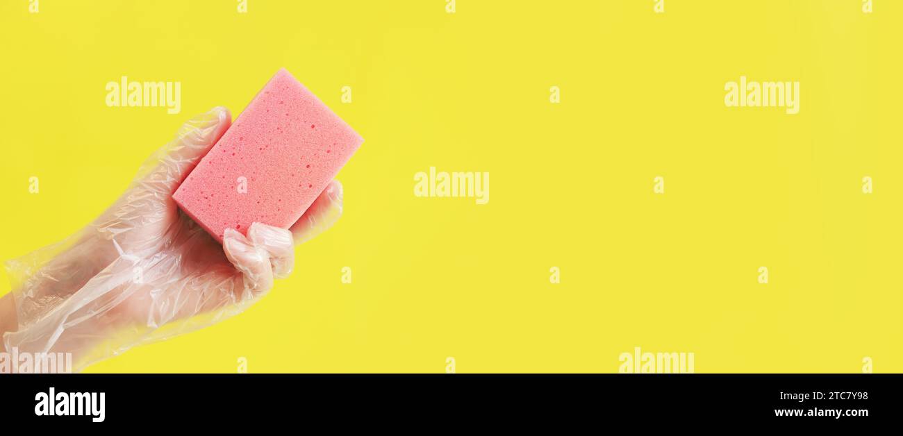 Ein rosafarbener Schwamm zum Geschirrwaschen in einer Frauenhand auf gelbem Hintergrund. Eine Hand in einem transparenten Einweghandschuh hält einen Schwamm für die Nassreinigung. Pro Stockfoto