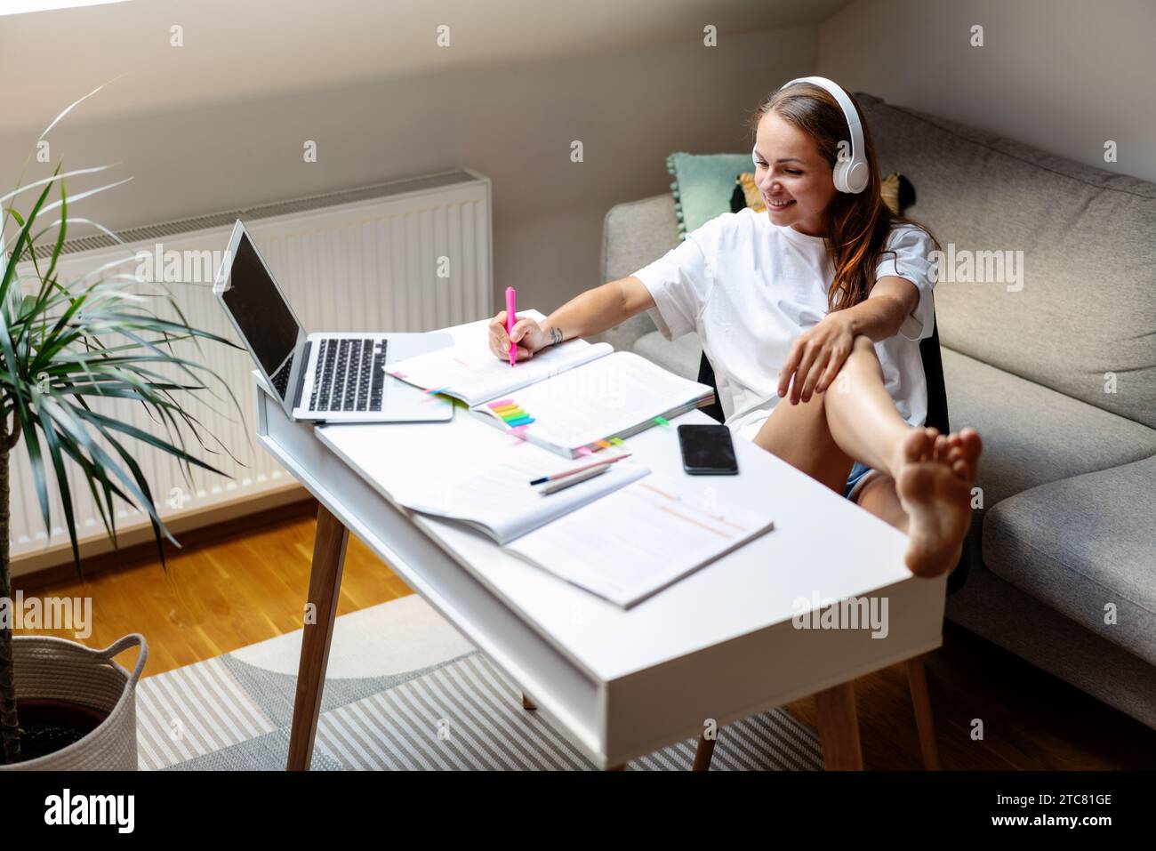 Häusliche Bildung. Attraktive brünette Frau in Kopfhörern, die ein Arbeitsbuch am Schreibtisch verwendet. Nutzung des Notebooks für E-Learning. Stockfoto