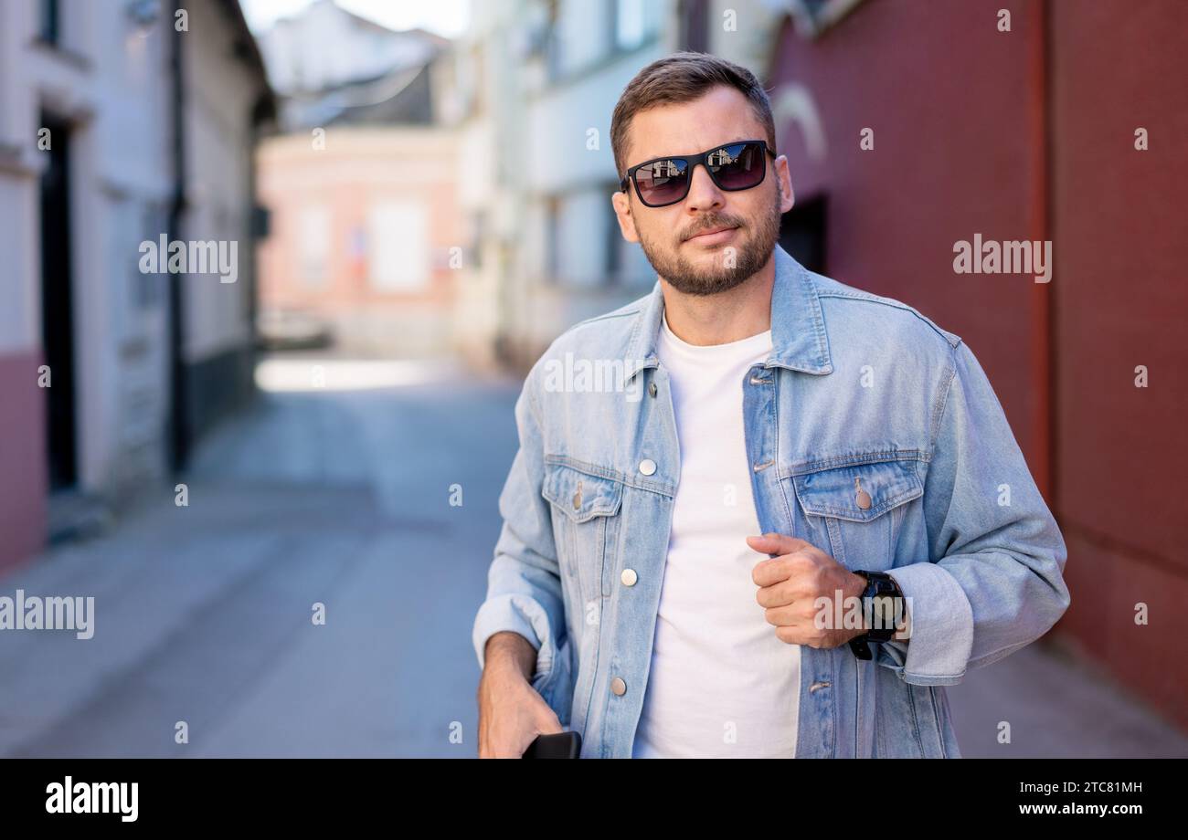 Junger erwachsener Mann mit Sonnenbrille und Jeansjacke posiert während er im Frühling auf der Straße steht. Stockfoto
