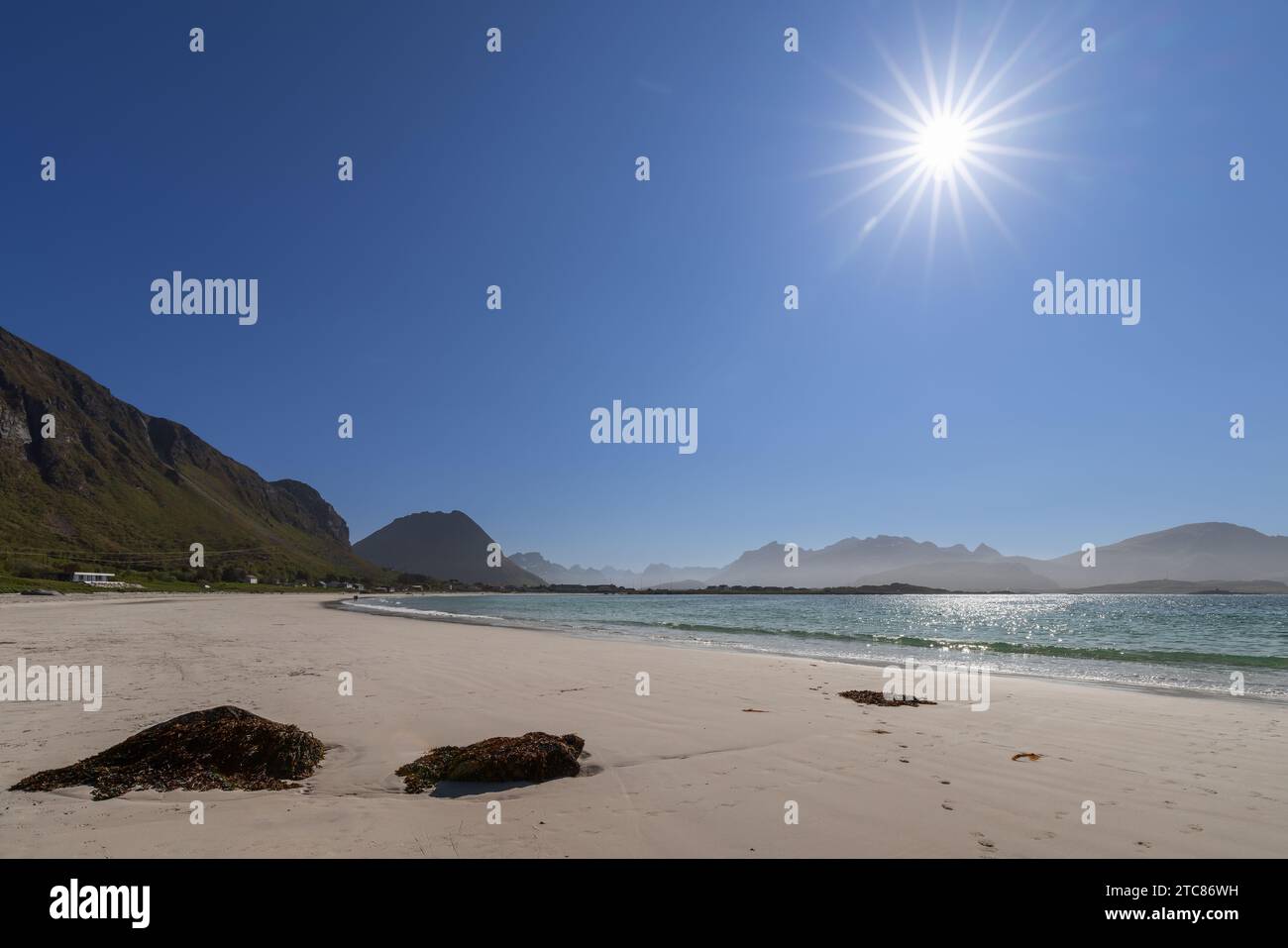 Strahlende Sonne über Jusnesvika Bay, Algen auf dem weißen Sand des Rambergstranda Strandes, mit den majestätischen Bergen der Insel Flakstadoya in d Stockfoto