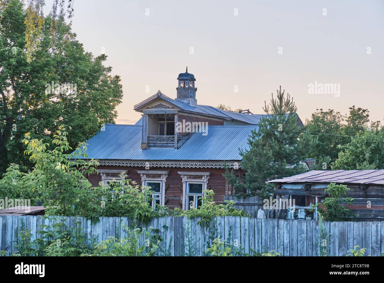 Jelabuga, Russland - 18. Juni 2023: Altes Holzhaus hinter verblasstem Zaun. Geschnitzte Holzleisten an den Fenstern, Dachbalkon, Türmchen mit verziertem Turm Stockfoto