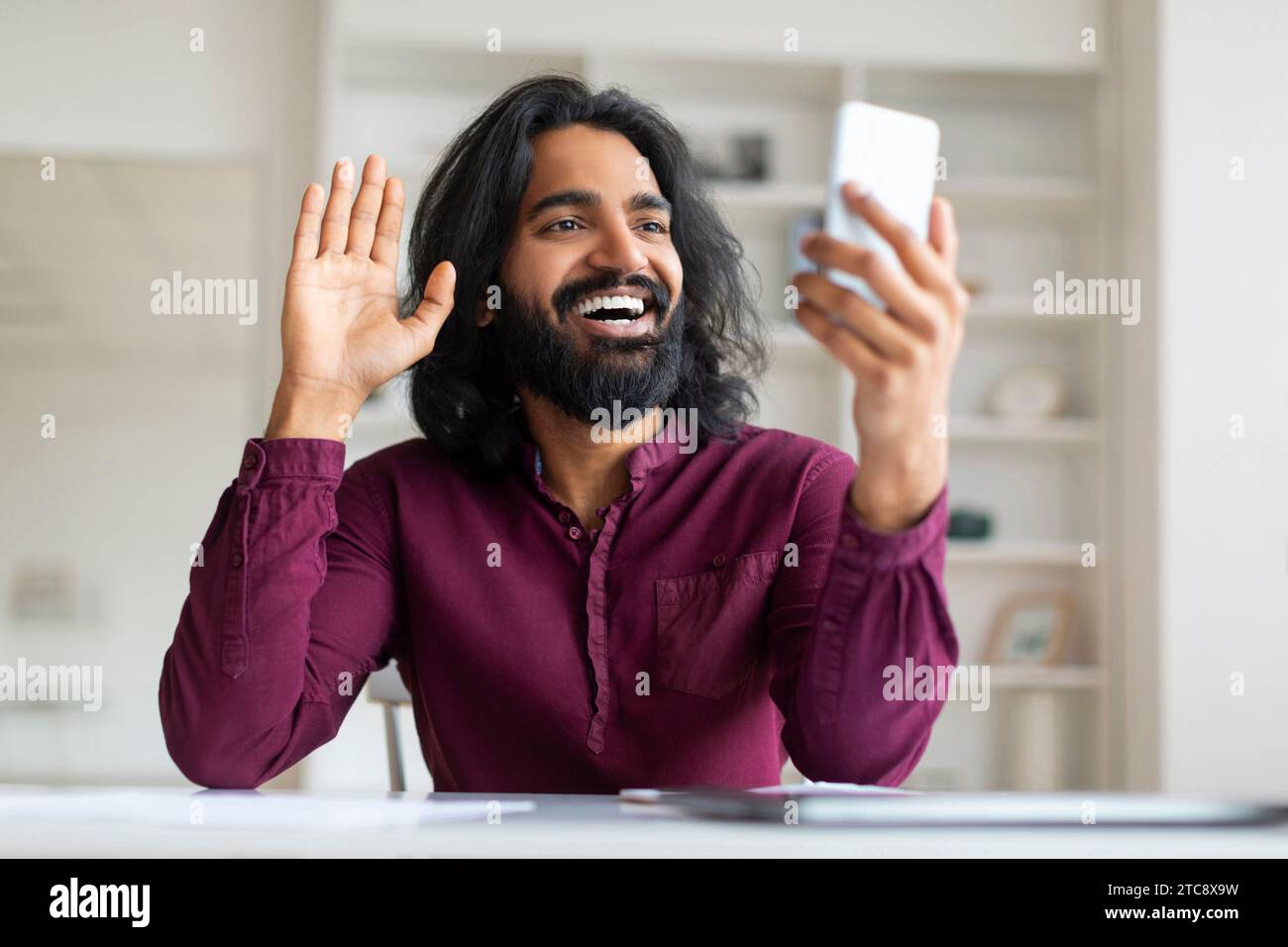 Lächelnder indischer Freelancer, der die Hand winkt, während er Videoanrufe auf dem Smartphone hat Stockfoto