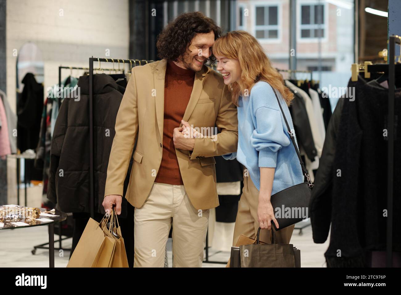 Fröhlicher junger Mann und Frau lachen, während sie in einer Boutique zwischen Regalen mit neuer saisonaler Modekollektion und kleiner Ausstellung stehen Stockfoto