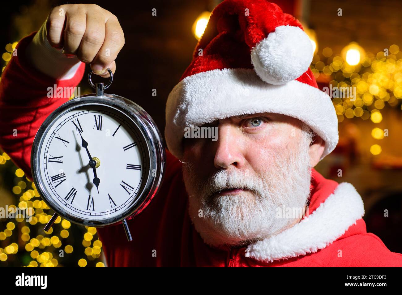 Ernster Weihnachtsmann mit weihnachtlicher Uhr. Zeit zum Feiern. Neujahr oder Weihnachten. Winterfeiertage. Bärtiger Mann mit Weihnachtsmann-Hut mit Stockfoto