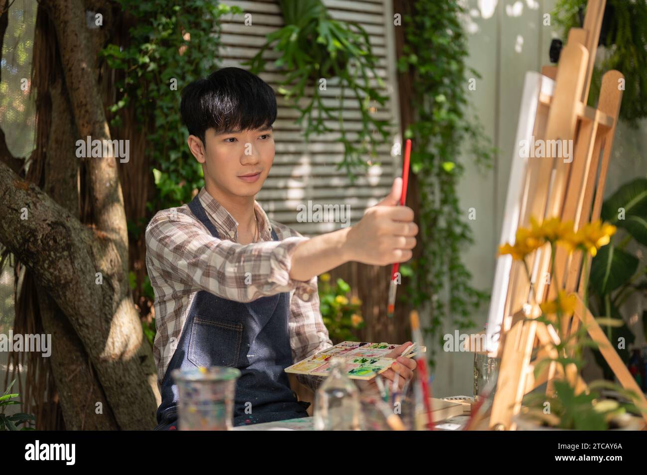 Ein fokussierter und gutaussehender junger asiatischer Mann in einer Schürze misst seine Zeichnung mit einem Pinsel auf einer Leinwand und macht ein Kunstwerk in seinem Garten. Stockfoto