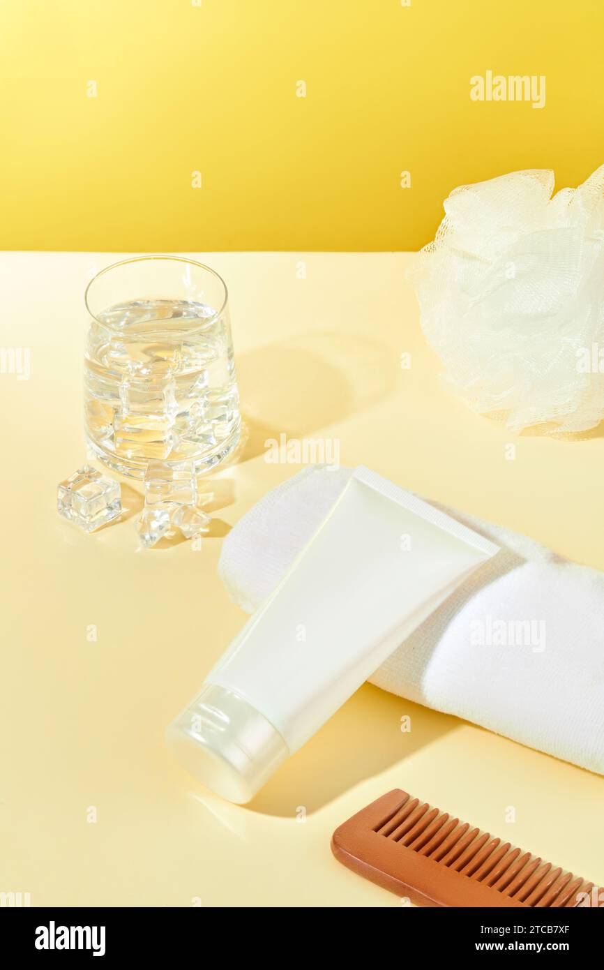 Ein Glas Wasser mit Eis, eine Tube Kosmetik ohne Marke, ein Badeschwamm, ein Kamm und ein Handtuch sind auf einem gelb-beigen Hintergrund ausgestellt. Werbung SK Stockfoto