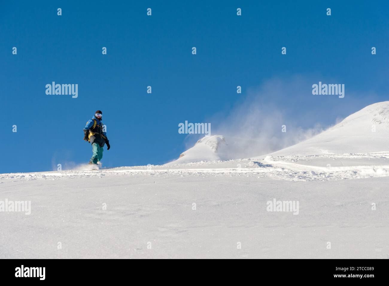 Ein Freeboard-Snowboarder in einer Skimaske und einem Rucksack läuft über die schneebedeckte Piste und hinterlässt einen Schneepulver am blauen Himmel Stockfoto