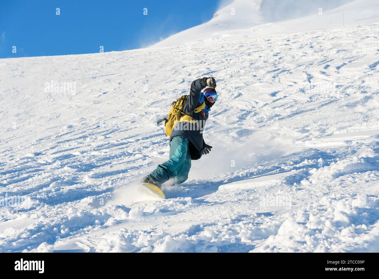 Ein Freeboard-Snowboarder in einer Skimaske und einem Rucksack läuft über die schneebedeckte Piste und hinterlässt einen Schneepulver am blauen Himmel Stockfoto