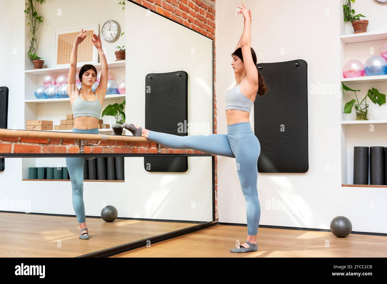 Ganzkörper-Seitenansicht einer jungen Ballerina in Aktivkleidung, die Dehnungsübungen mit erhobenen Armen vor dem Spiegel im Fitnessstudio macht Stockfoto