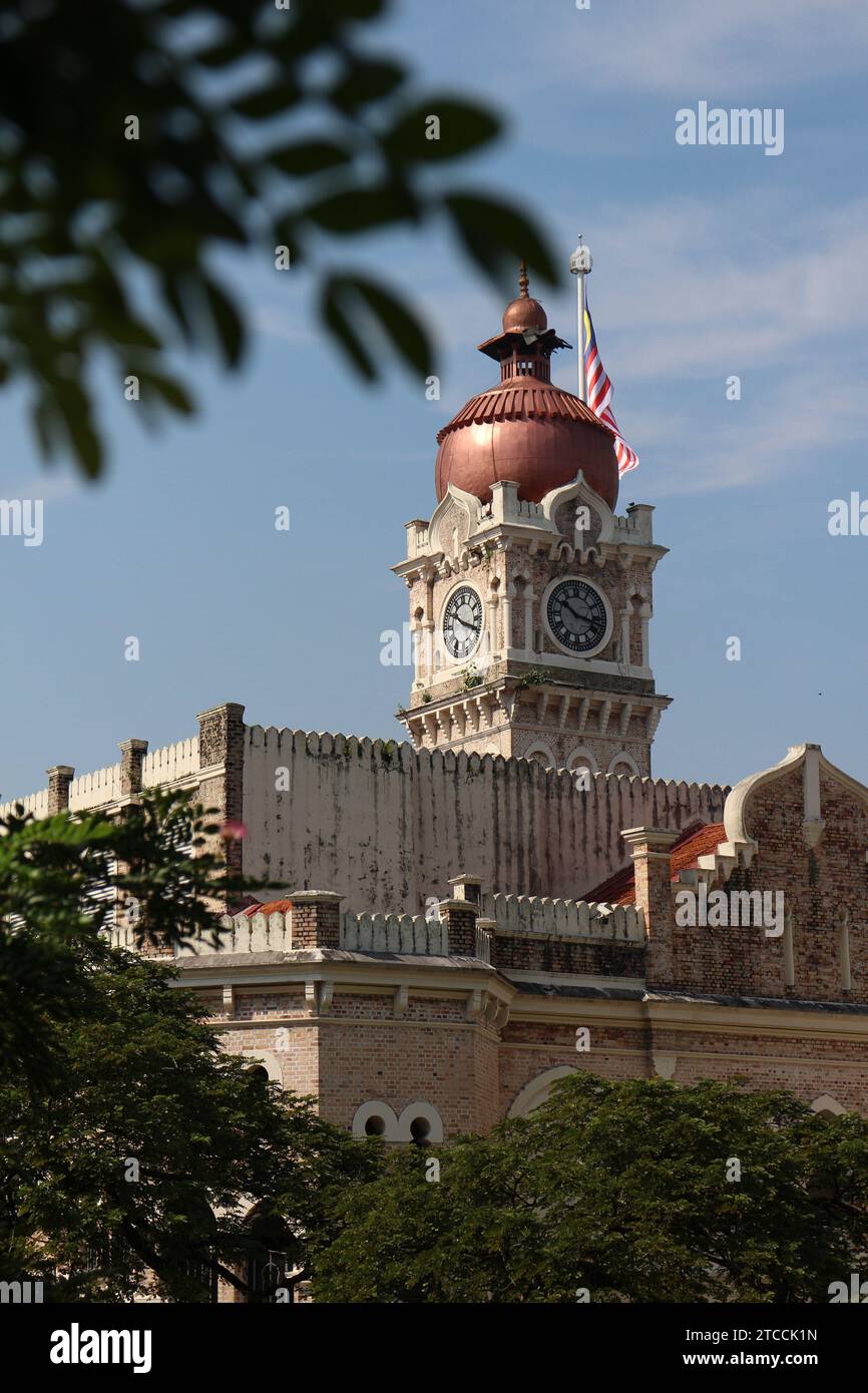 Ein beeindruckender Glockenturm hebt sich vor der Kulisse eines alten Gebäudes und üppiger Grünflächen hervor Stockfoto