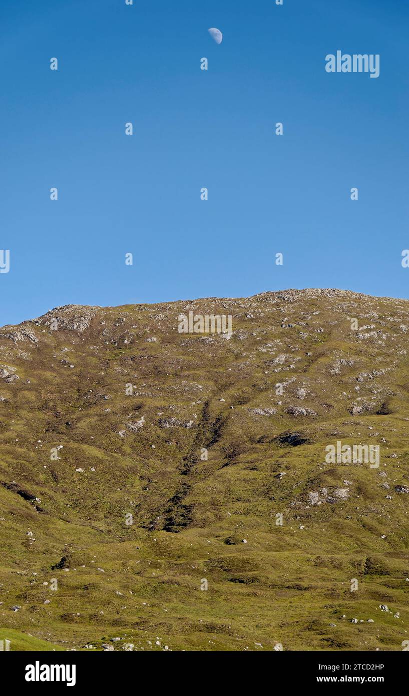 Der Mond, der in einem blauen Himmel über einem zerklüfteten Hügel in North Lochs auf der Isle of Lewis in den Äußeren Hebriden Schottlands aufsteigt. Stockfoto