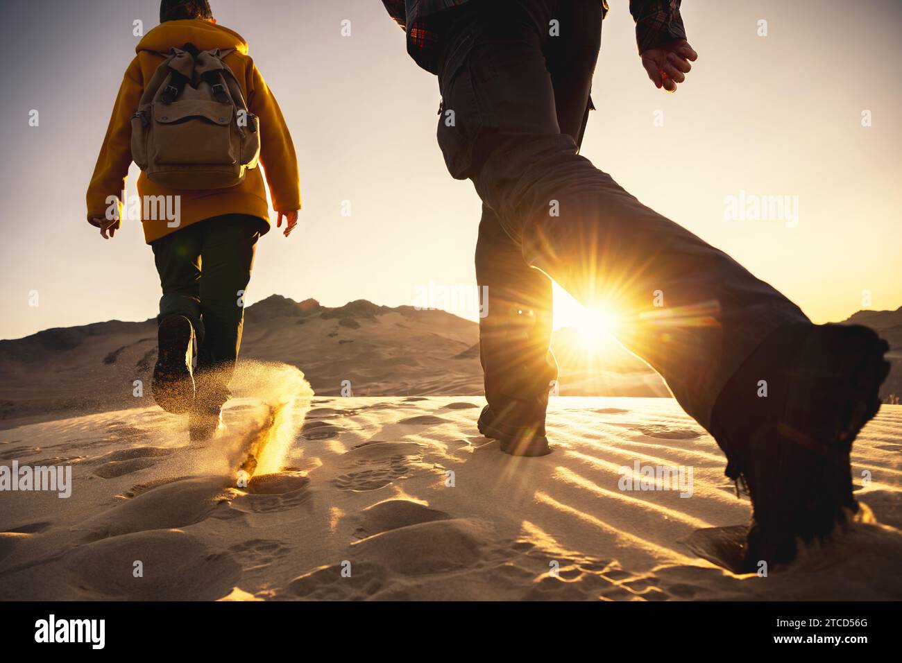 Zwei Wanderer mit Rucksäcken spazieren durch Wüstendünen bei Sonnenuntergang. Nahaufnahme von Wanderschuhen. Wanderkonzept Stockfoto