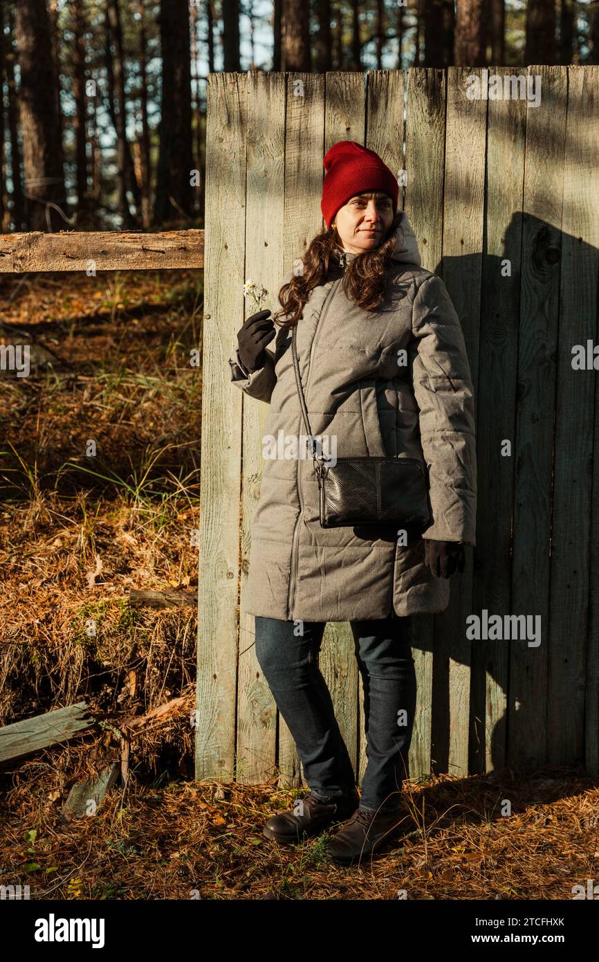 Eine modische Frau mittleren Alters steht selbstbewusst in ihrem Herbstensemble neben einem alten, schäbigen Zaun und strahlt Wärme und Eleganz auf einem entzückenden Su aus Stockfoto