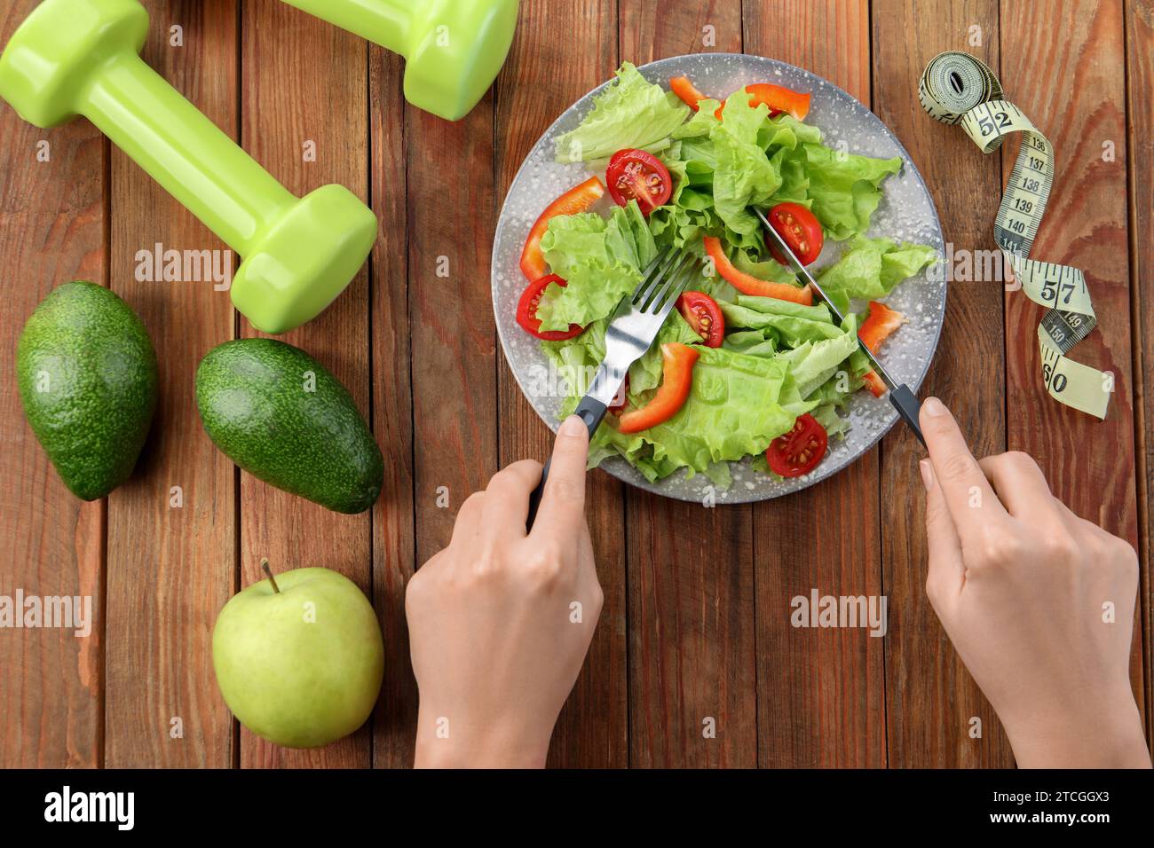 Gesunde Ernährung. Frau isst Salat am Holztisch mit Maßband und Kurzhanteln, Blick von oben Stockfoto