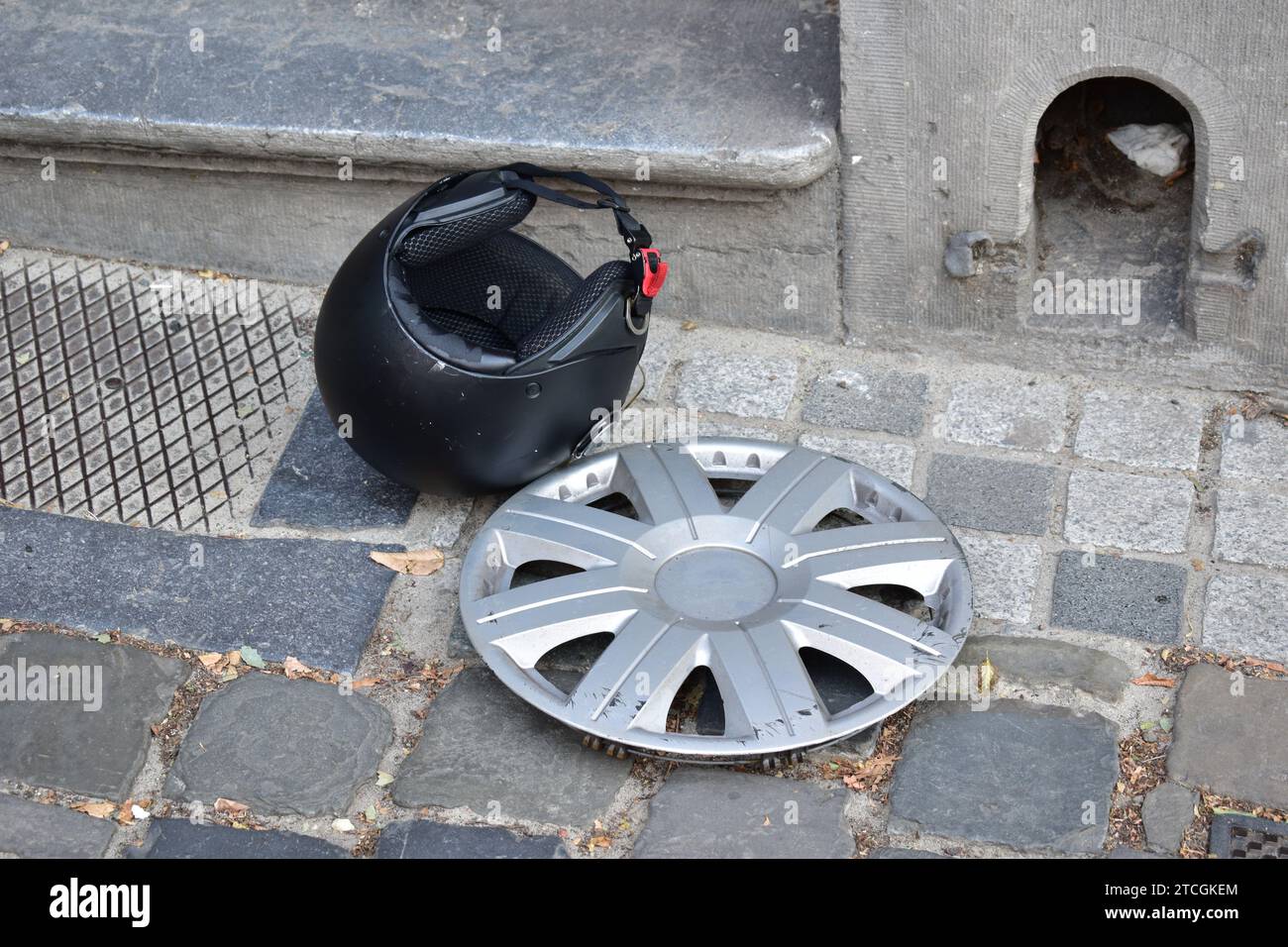 Eine kratzende graue Kunststoffabdeckung und ein mattschwarzer Motorradhelm liegen auf einem Kopfsteinpflaster im Stadtzentrum von Brüssel Stockfoto