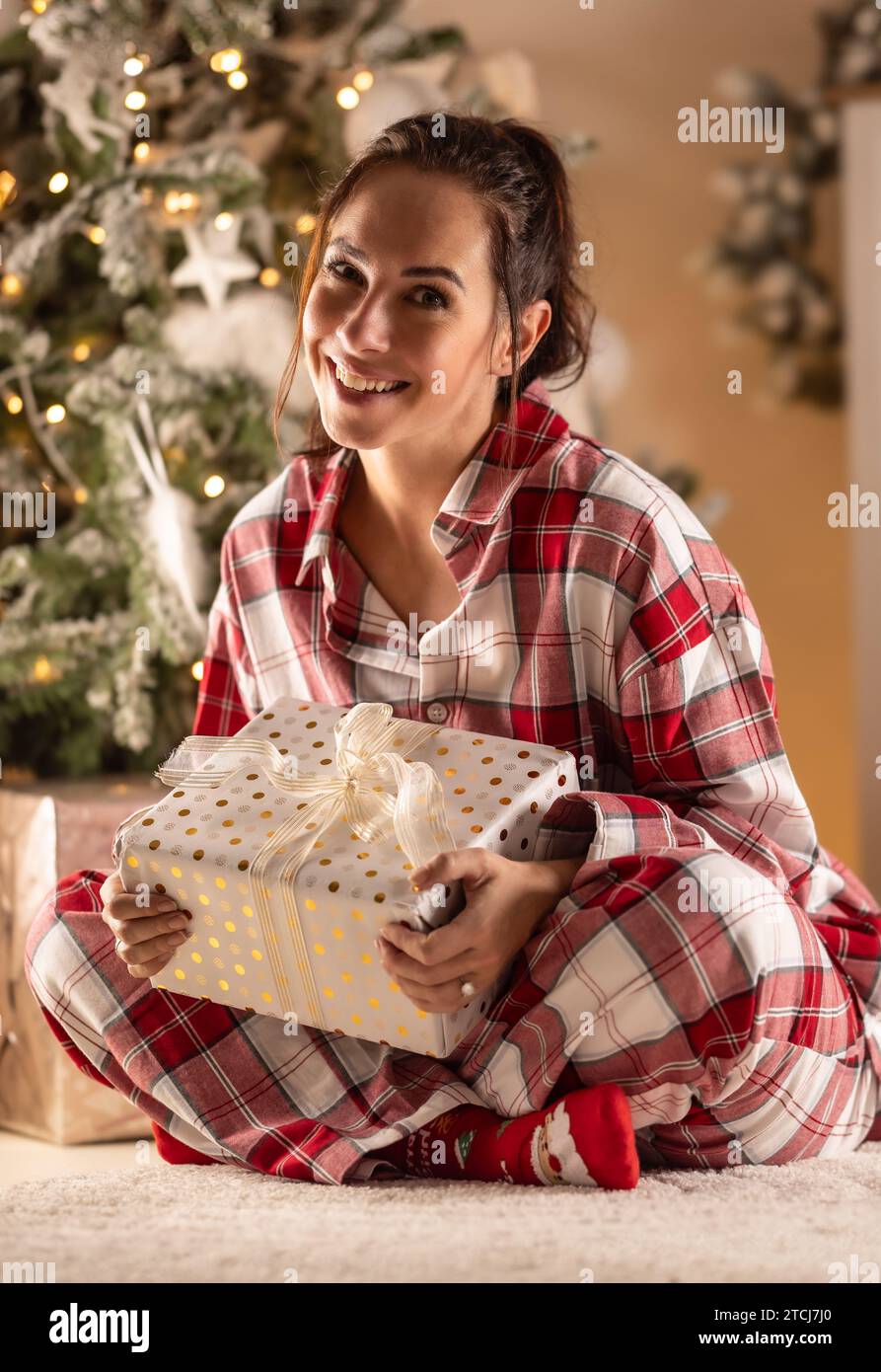 Eine süße Brünette sitzt neben dem Weihnachtsbaum und hält ihr Weihnachtsgeschenk. Stockfoto