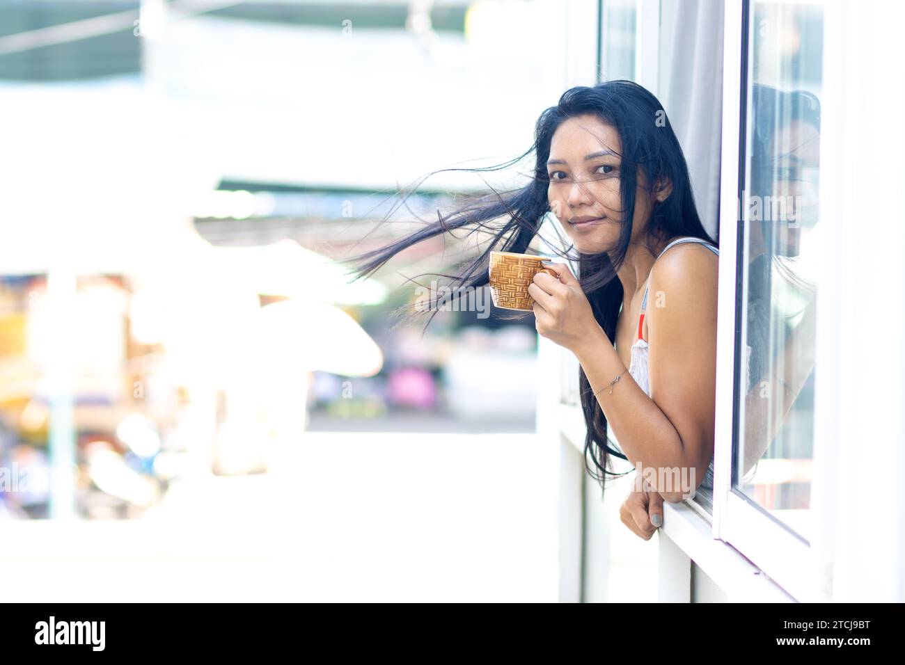 Eine Frau im Sommerkleid hält einen Becher in einem offenen Fenster eines Wohnhauses, während der Wind ihr die Haare weht Stockfoto