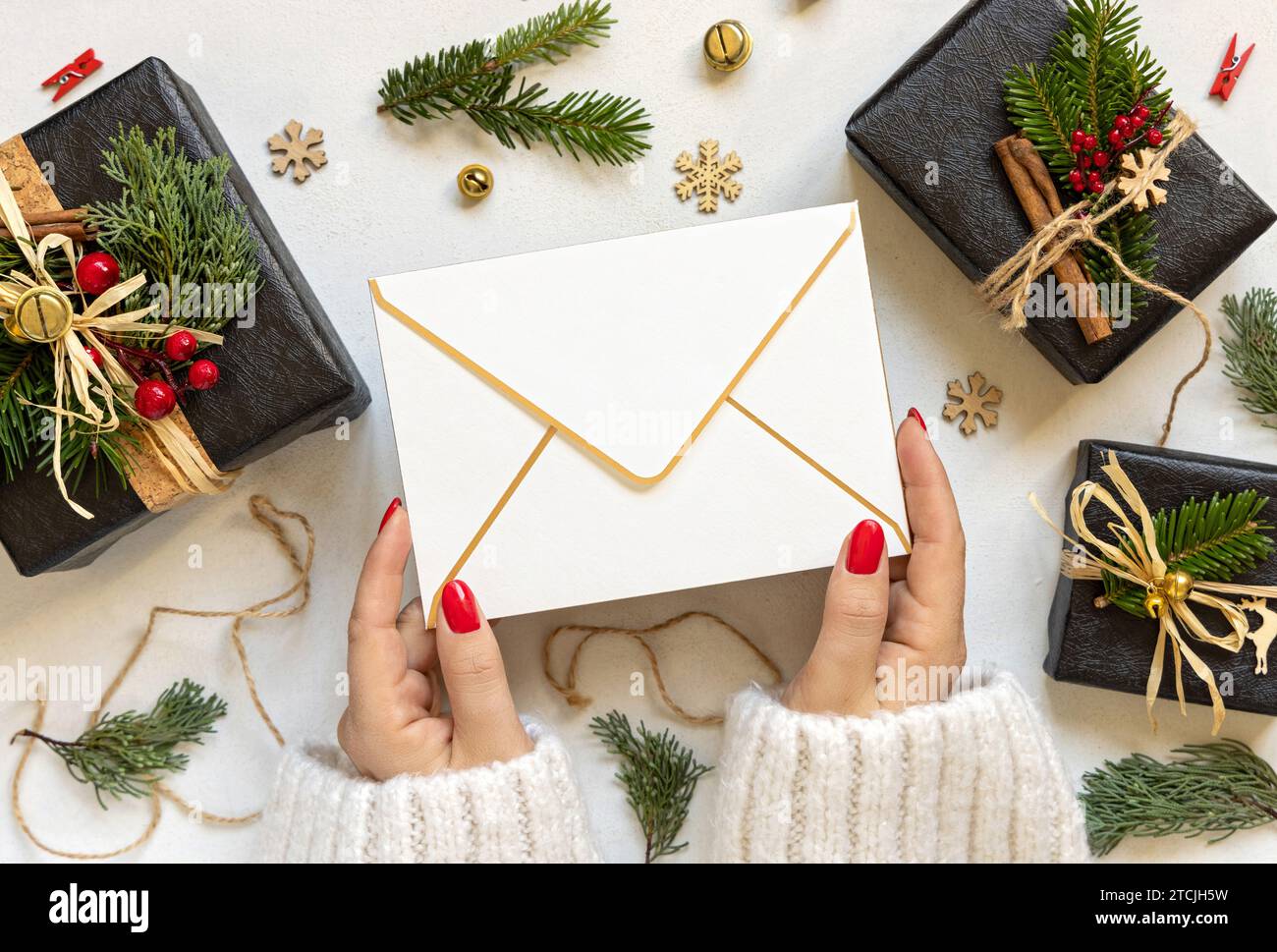 Hände mit Umschlag zwischen Weihnachtsdekor, schwarze Geschenkboxen und grüne Tannenzweige von oben. Leeres Umschlagmodell, flache Ladefläche, Kopierraum. Weihnachten Stockfoto