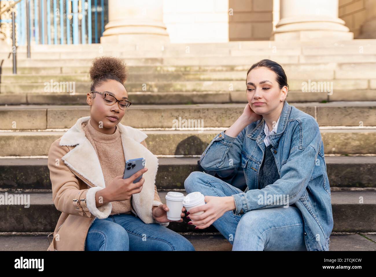 Zwei Studentinnen in Jeansjacke reden miteinander, am Telefon, lachen und trinken Kaffee auf den Treppen in der Stadt. Konzept der digitalen Teilung Stockfoto