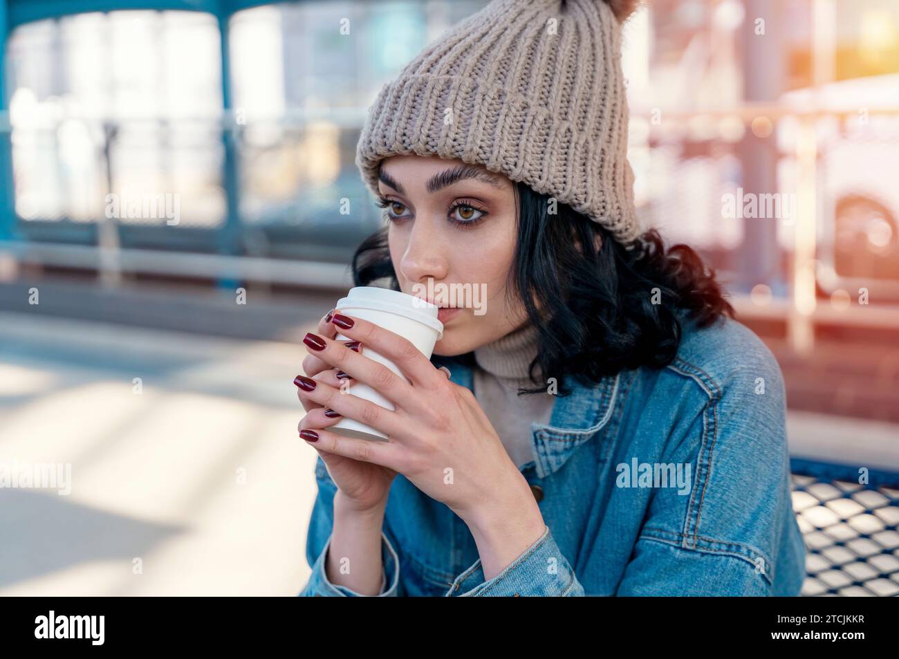 Eine junge Frau in einer Jeansjacke trinkt Kaffee und wartet auf eine Straßenbahn, Bus an der Haltestelle Lifestyle Foto Stockfoto