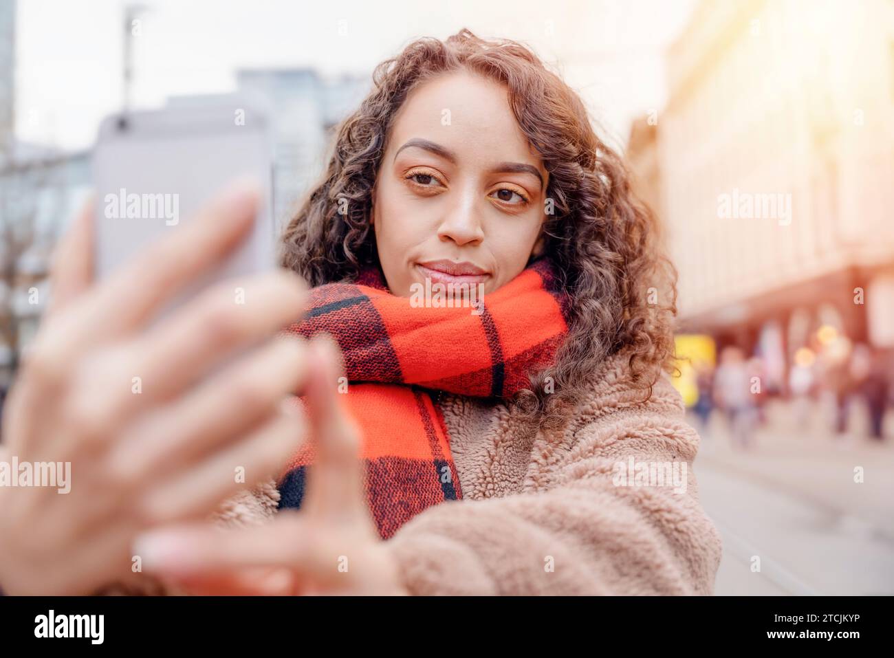 Eine fröhliche Frau, die Selfies auf einem Smartphone macht, Eine lockige brünette Frau in einer Jacke und einem orangefarbenen Schal, die lächelt und durch Europa reist. Stockfoto