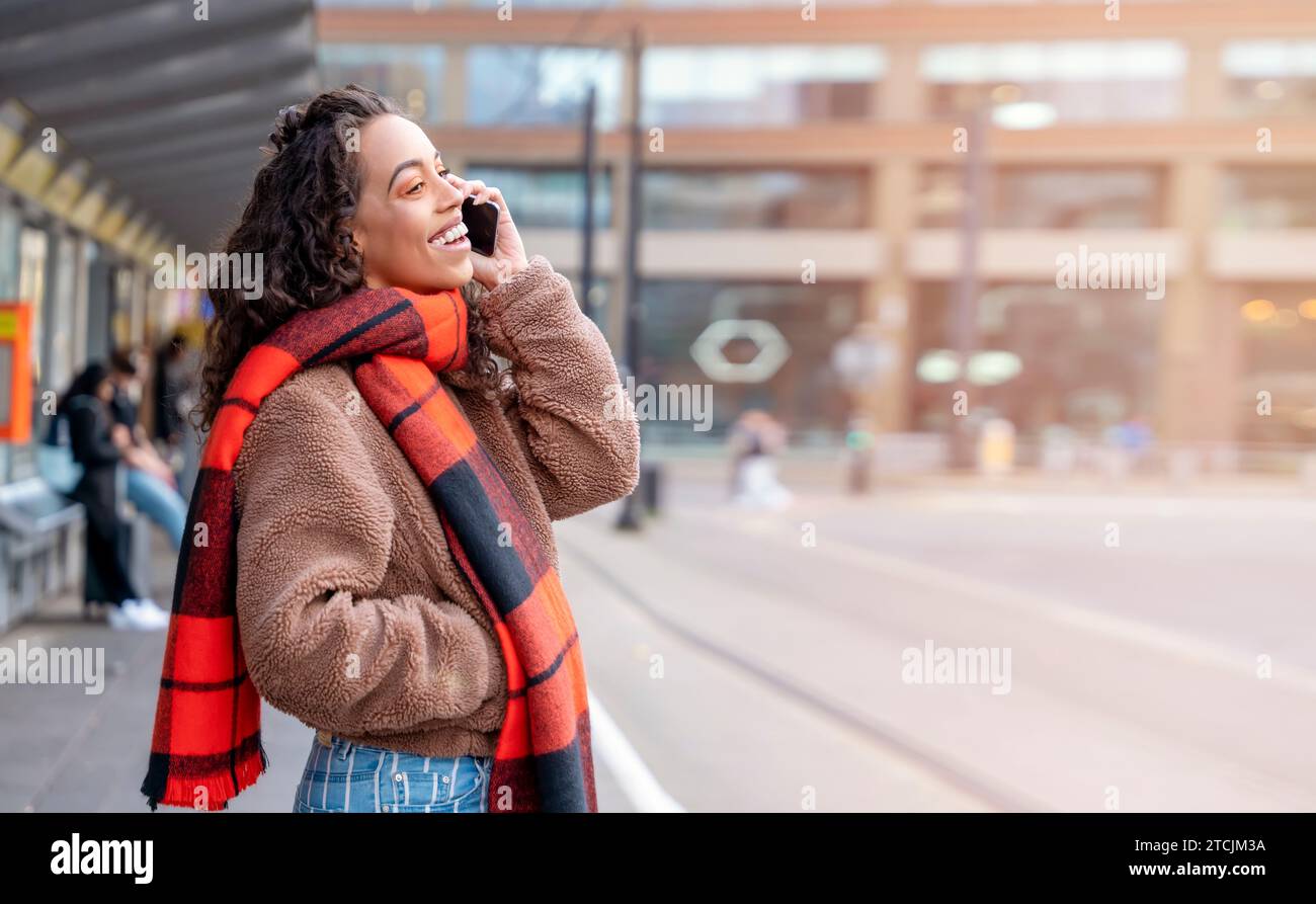 Junge Frau in Jacke und orangefarbenem Schal, die an der Haltestelle auf eine Straßenbahn wartet und auf einem Lifestyle-Foto spricht Stockfoto