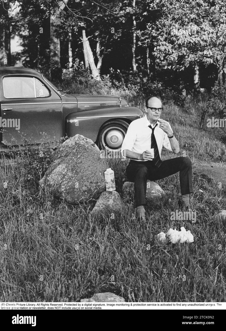 In den 1970er Jahren Man sieht, dass ein Mann nach der Straße angehalten hat, offenbar hungrig. Er saß auf einem Felsen und isst ein Sandwich und trinkt Saft, die Verpackung ist daneben. Er trägt Hemd und Krawatte. Der Wagen ein schwedisches Automodell Volvo PV 26 Juli 1971 Stockfoto