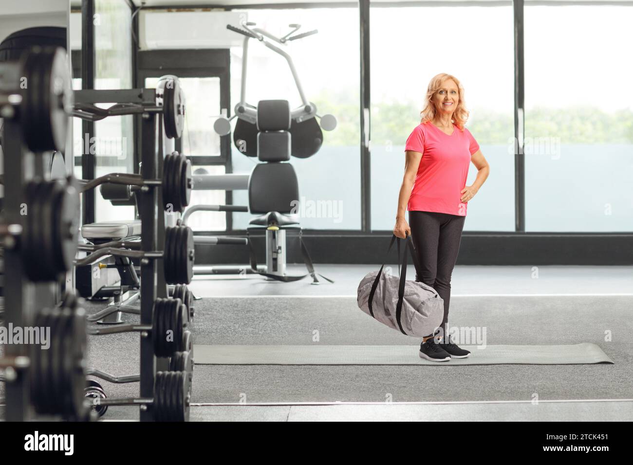 Lächelnde reife Frau in Sportbekleidung, die eine Sporttasche in einem Fitnesscenter hält Stockfoto