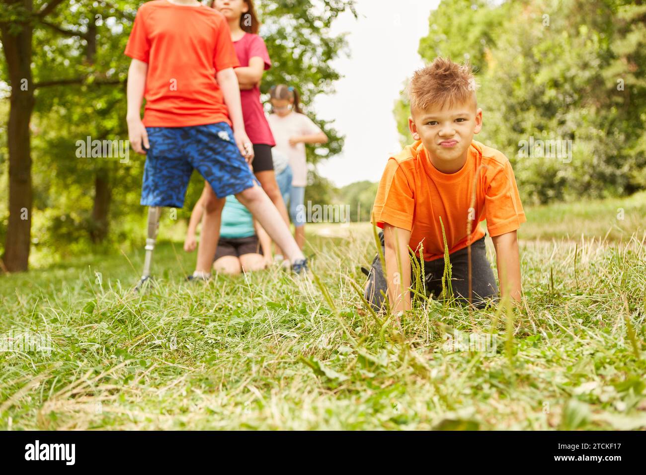 Porträt eines Jungen mit orangefarbenem T-Shirt, der auf Gras kniet, mit Freunden, die im Hintergrund im Park stehen Stockfoto