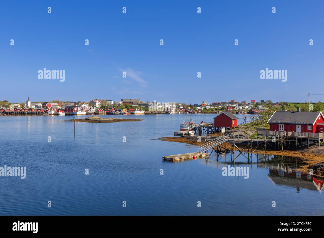 Ruhige Aussicht auf das reine Dorf in Lofoten, Norwegen, mit traditionellen roten Rrorbu-Hütten, ruhigem blauem Wasser und Fischerbooten unter klarem Himmel Stockfoto