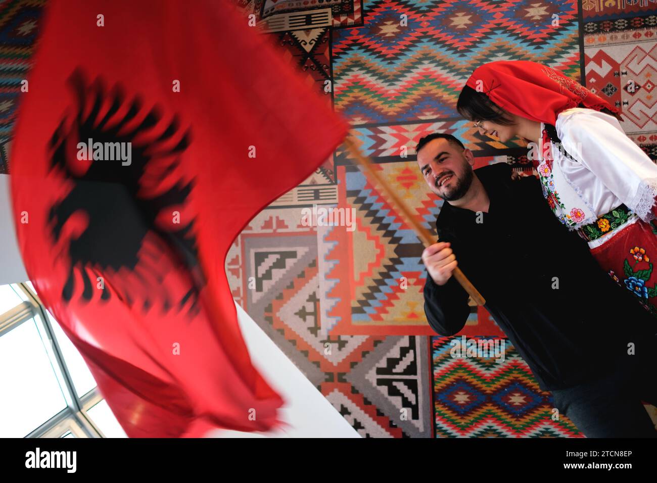 Tirana, Albanien – 28. November: Eine Frau und ein Mann in traditioneller albanischer Kleidung, die mit einer Flagge vor traditionellen Mustern posieren Stockfoto