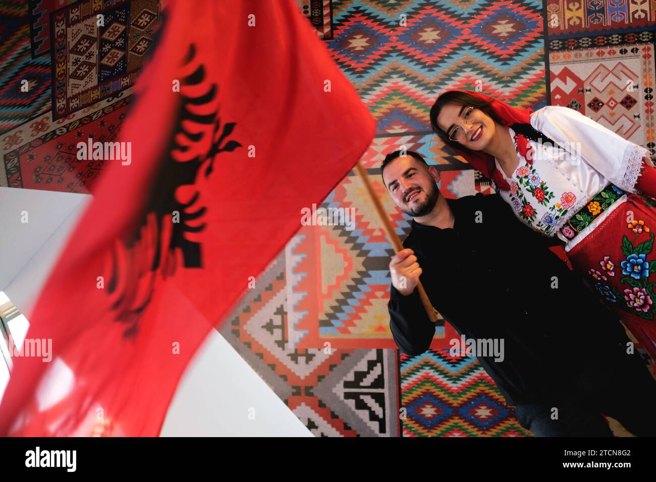 Tirana, Albanien – 28. November: Eine Frau und ein Mann in traditioneller albanischer Kleidung, die mit einer Flagge vor traditionellen Mustern posieren Stockfoto