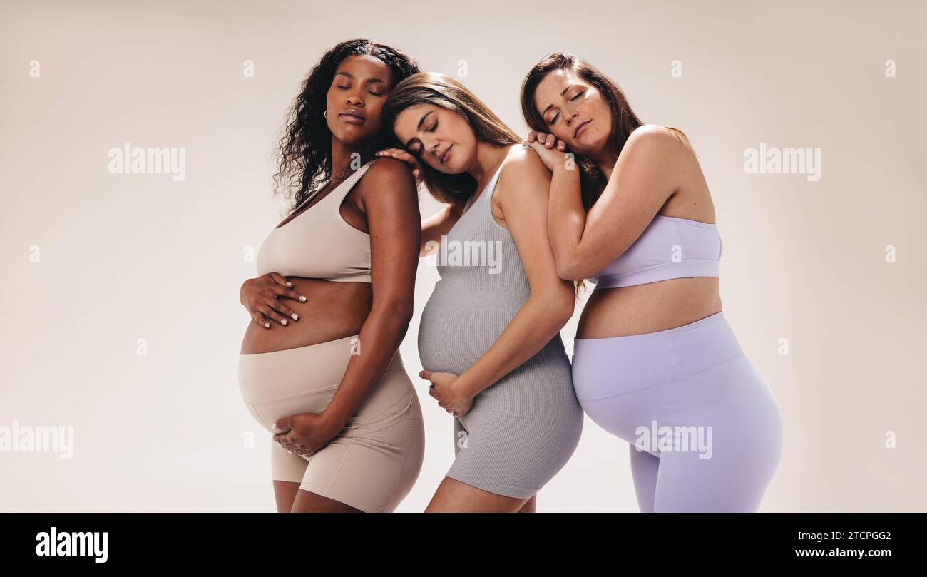 Multikulturelle schwangere Frauen, die sich in einem Fitnessstudio aneinander lehnen und stehen. Weibliche Freunde, die einen Yoga-Kurs benutzen, um sich während der Zeit gegenseitig zu unterstützen Stockfoto