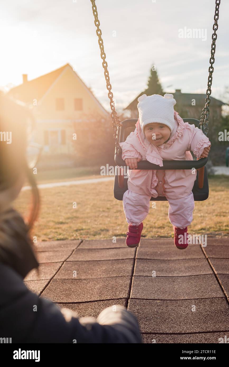 Adorable Baby Mädchen mit grossen schönen Augen und einem beanie Spaß auf einer Schaukel Fahrt auf einem Spielplatz in einem sonnigen Park Stockfoto