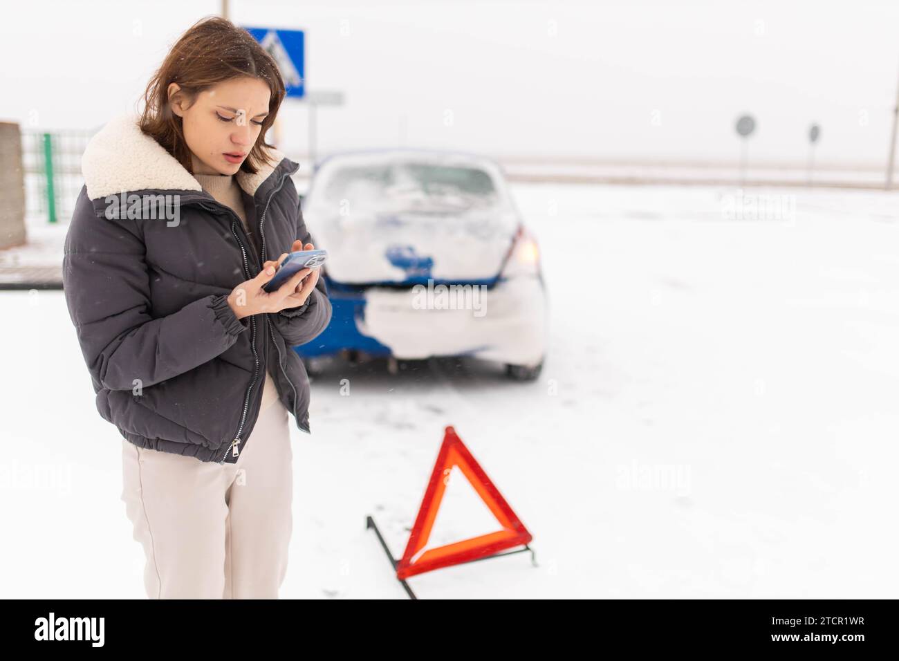 Eine junge Fahrerin, die ihr Auto auf einer rutschigen Winterstraße im Schnee zertrümmert hat, ruft einen Abschleppwagen mit einem Notdreieck neben ihr in der Nähe des Autos an Stockfoto