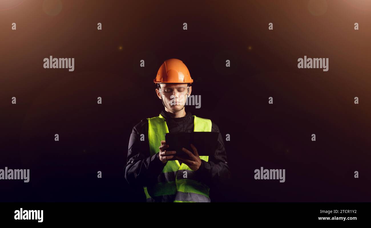 Engineering Consulting, ein männlicher Bauarbeiter in einem Schutzhelm auf dunklem Hintergrund benutzt ein Tablet. Geschäftsprozessmanagement und Bauinspektor mit Stockfoto