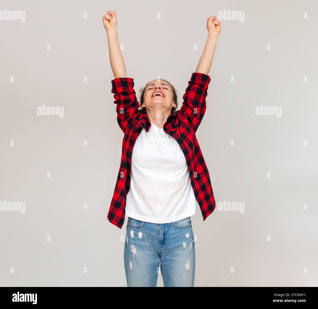 Frau in legeren Kleidern, die vor einem schlichten Hintergrund mit erhobenen Armen steht. Freuen Sie sich, feiern Sie den Sieg. Stockfoto