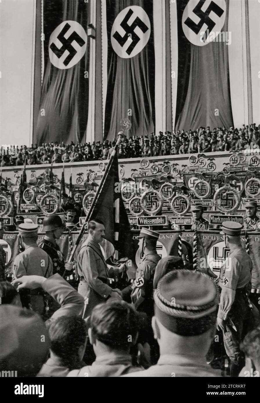 Während des Reichsparteitags 1934 wird Adolf Hitler in einem bedeutenden Moment gefangen genommen und schüttelt bei der Zeremonie der „Weihe der Standards“ die Hand eines Soldaten. Dieses Ereignis symbolisierte die Einheit und Loyalität der Streitkräfte gegenüber dem Nazi-Regime. Hitlers persönliches Engagement, wie durch den Handschlag belegt, unterstreicht seine zentrale Rolle bei diesen Machtdemonstrationen und der bewussten Kultivierung einer Bindung zwischen der Partei und dem Militär. Stockfoto
