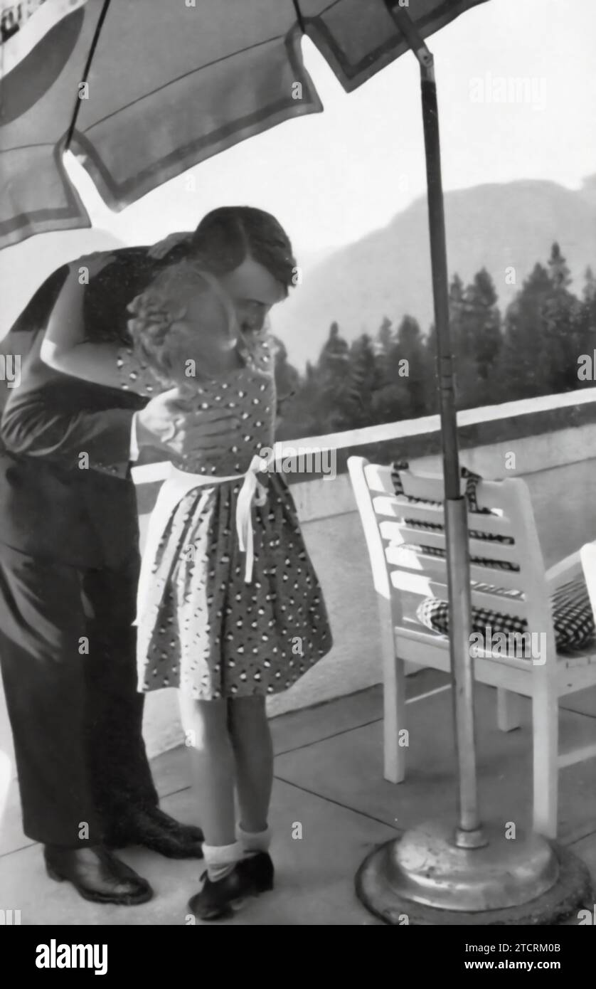 Adolf Hitler wird in einem ruhigen Moment in Obersalzberg gefangen genommen, mit einem jungen Mädchen, das ihn umarmt. Dieses Bild auf einer Terrasse mit malerischer Bergkulisse zeigt eine sanfte und mitfühlende Seite Hitlers, ein krasser Kontrast zu seinem üblichen autoritativen Verhalten. Stockfoto