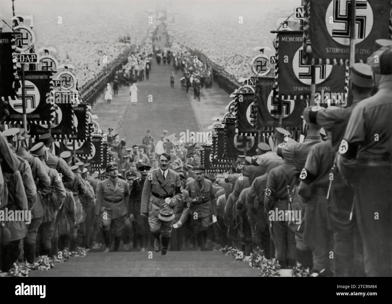 Adolf Hitler wird bei einer massiven Kundgebung der NSDAP in Bückeberg 1934 die Treppe hinaufsteigen sehen. Diese Veranstaltung, Teil des jährlichen Erntefestes, war ein bedeutendes Propagandawerkzeug, das die Fähigkeit des Regimes demonstrierte, große Menschenmengen unter seiner Ideologie zu mobilisieren und zu vereinen. Die Rallye 1934 war besonders für ihren großen Umfang und die orchestrierte Darstellung von Loyalität und Einheit innerhalb der Partei bekannt. Stockfoto
