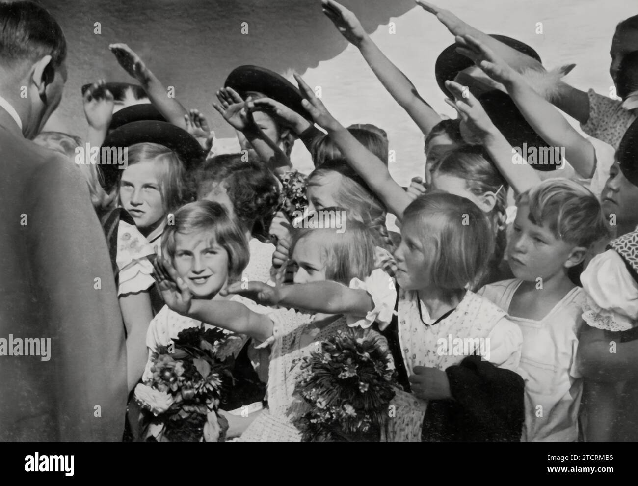 In der Kinderhände wird Adolf Hitler gesehen, wie er Kinder grüßt und Blumen von ihnen erhält. Dieses Bild ist ein ergreifendes Beispiel dafür, wie das Nazi-Regime orchestrierte Interaktionen mit Jugendlichen nutzte, um ein öffentliches Bild von Wohlwollen und Zugänglichkeit zu pflegen. Der Akt der Annahme von Blumen von Kindern war ein verbreitetes Propagandawerkzeug, das Hitlers Image weicher machen und die Idee eines pflegenden, väterlichen Führers fördern sollte, der eng mit der jüngeren Generation verbunden ist. Stockfoto